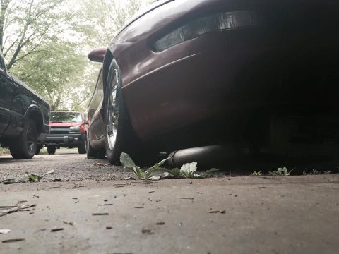 1995 Ford Probe - Low-angle shot of the rear of a dark red car, possibly a 1995 Ford Probe, showing its exhaust pipe and wheel. A black pickup truck and a re…