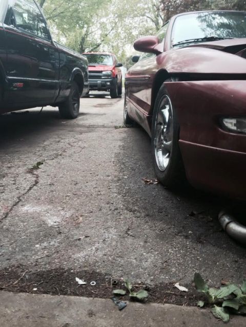 1995 Ford Probe - A low-angle shot shows the side of a red coupe parked on an asphalt driveway next to a black pickup truck. Another red vehicle is visible i…