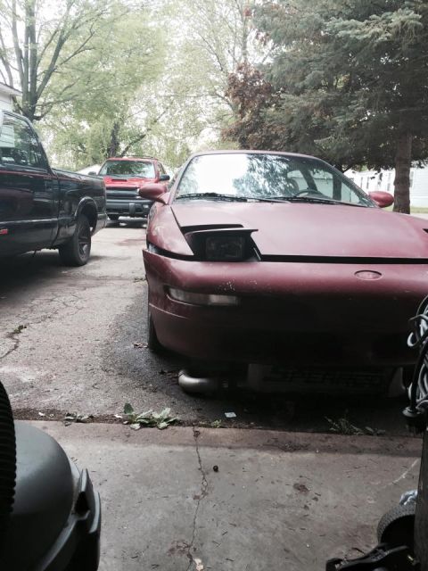 1995 Ford Probe - A maroon Ford Probe is parked in a driveway next to a black pickup truck. The front of the Probe is visible, showing its pop-up headlights…