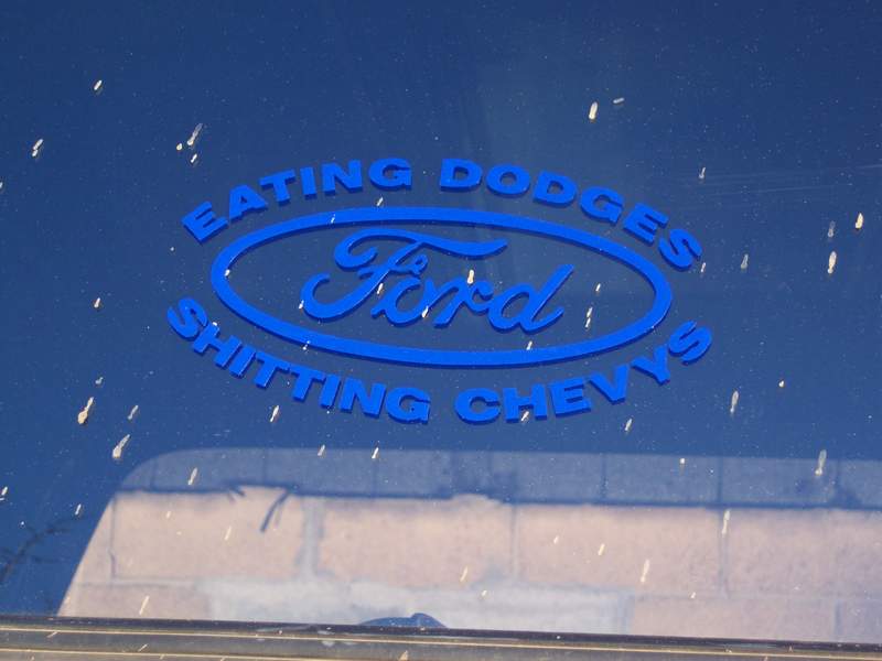 Blue vehicle window with a blue oval Ford logo sticker that says "EATING DODGES SHITTING CHEVY'S"