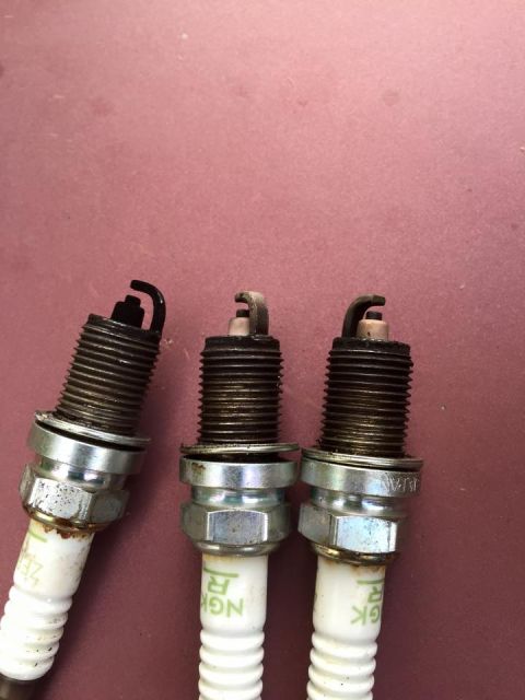 1995 Ford Probe - Three NGK spark plugs are shown against a reddish-brown background. The spark plugs have metal threaded bases and ceramic insulators with v…