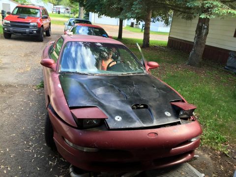 1995 Ford Probe - A maroon 1995 Ford Probe with a black hood sits parked on a gravel driveway next to a red SUV. The car's pop-up headlights are raised, and…