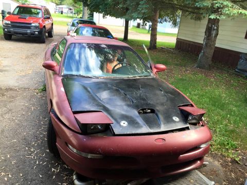 1995 Ford Probe - A maroon Ford Probe with a black hood is parked in a driveway with a red SUV in the background. The car's headlights are popped up, and a p…