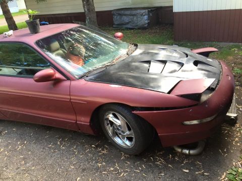 1995 Ford Probe - A maroon 1995 Ford Probe is parked outdoors with a modified black hood featuring vents. The car's front end is visible, showing pop-up head…