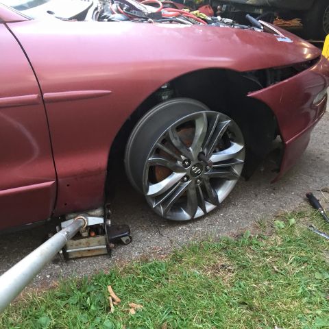 1995 Ford Probe - A maroon coupe is jacked up with the front wheel removed, revealing the brake rotor. A car jack is positioned under the frame, and tools ar…