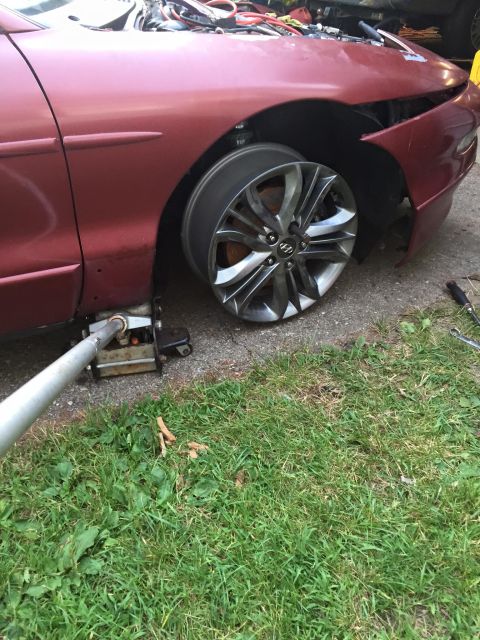 1995 Ford Probe - A red car is jacked up with the front passenger wheel removed, revealing the brake rotor. A silver jack handle is visible in the foreground.