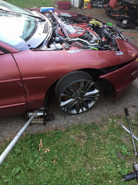 1995 Ford Probe - A red coupe is jacked up with the front passenger wheel removed, exposing the engine bay. Tools are laid out on the grass next to the vehic…