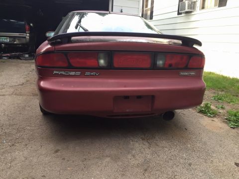 1995 Ford Probe - The rear of a red 1995 Ford Probe GT is shown parked on a gravel driveway in front of a garage. The car has a spoiler, a GT badge on the ta…