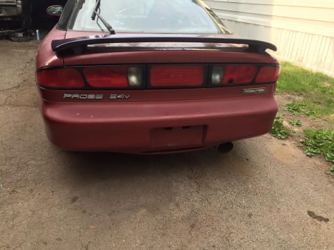 1995 Ford Probe - The rear of a red 1995 Ford Probe GT is shown parked on a gravel driveway next to a building. Visible badging includes 'PROBE 24V' and 'MAZ…