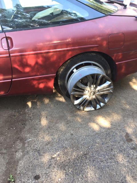 1995 Ford Probe - The rear passenger side of a red 1995 Ford Probe is shown with a flat tire and a detached silver alloy wheel resting on the ground next to…