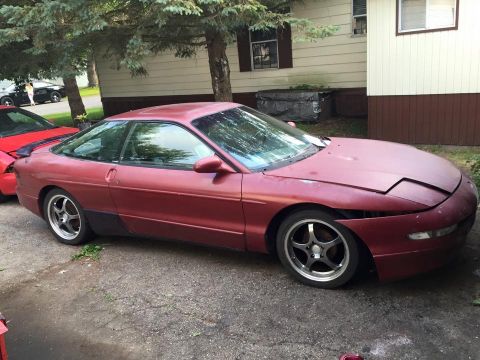 1995 Ford Probe - A maroon Ford Probe is parked on a gravel driveway next to a red car. The front passenger side fender is missing, exposing the wheel well.