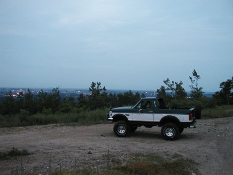 1994 Ford Bronco - A lifted, two-tone pickup truck sits on a dirt path with a city skyline visible in the distance under a cloudy sky.