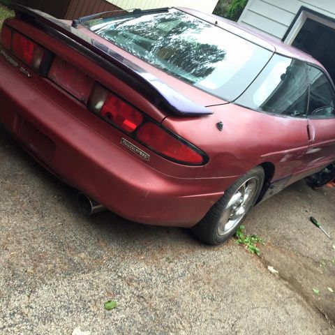 1995 Ford Probe - The rear passenger side of a red 1995 Ford Probe is shown parked on a concrete driveway. The car has a dark spoiler and a visible exhaust p…