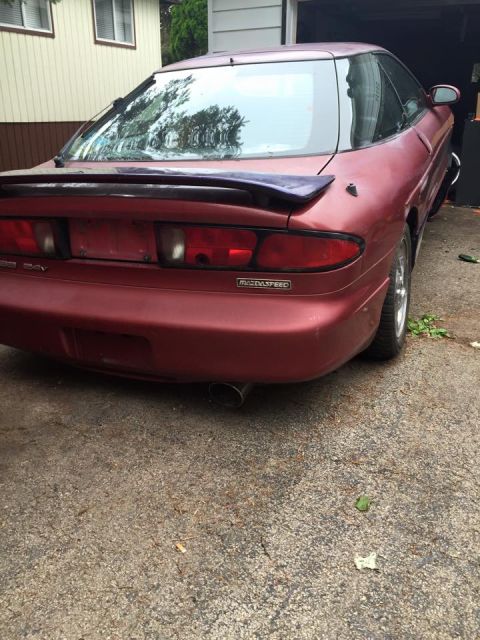 1995 Ford Probe - The rear driver's side quarter view of a red MazdaSpeed Ford Probe is shown parked on a concrete driveway. The car features a dark purple r…