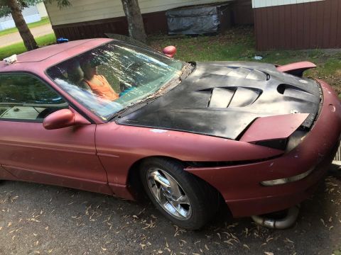 1995 Ford Probe - A maroon 1995 Ford Probe is parked outdoors with a black aftermarket hood. A person wearing an orange tank top is visible through the winds…