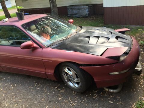 1995 Ford Probe - A maroon 1995 Ford Probe with a black aftermarket hood sits parked on a gravel driveway. The car has pop-up headlights and a plant in a pot…