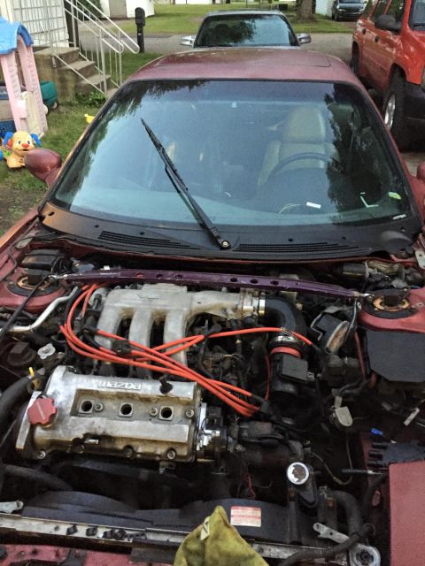 1995 Ford Probe - The engine bay of a red car is shown, with the word 'MAZDA' clearly visible on the engine block. Another vehicle is parked behind it.