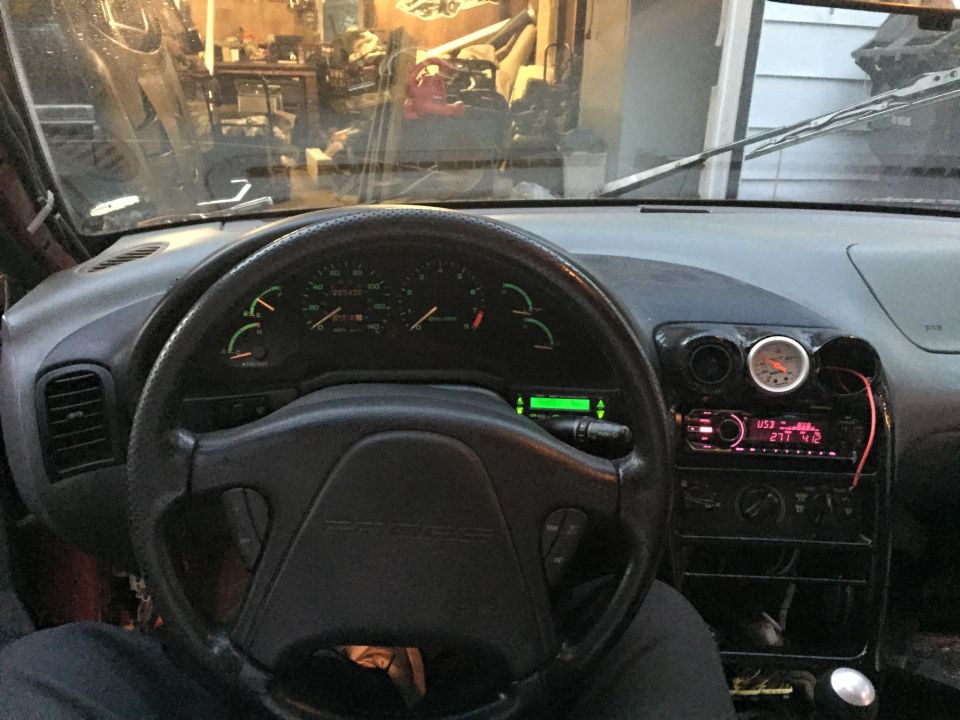 Car dashboard with steering wheel, gauges, and radio. Aftermarket gauge above the radio