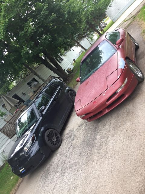 1995 Ford Probe - A red sports car with pop-up headlights is parked next to a black SUV on a paved surface.