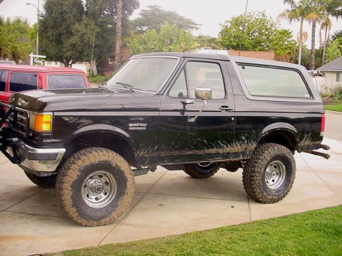 1988 Ford Bronco - A black lifted Ford Bronco with mud splatters sits on a paved driveway, with a red SUV visible behind it.