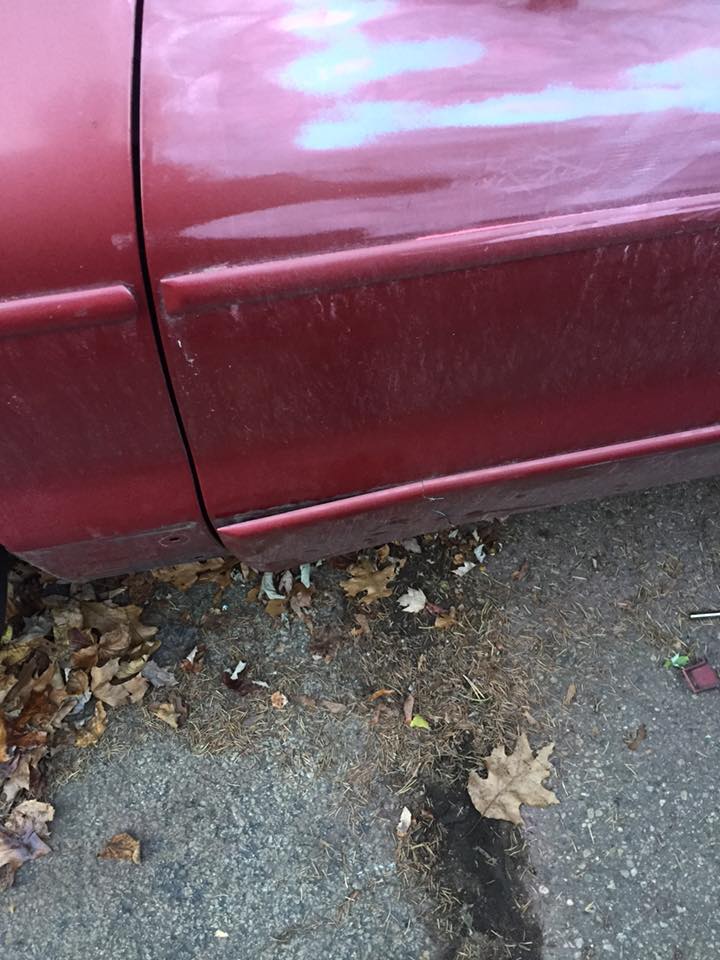 Red car door with visible panel gap and some dirt. Fallen leaves are on the ground below