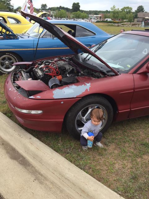1995 Ford Probe - A young child squats beside a red 1995 Ford Probe with its hood open, parked at an outdoor car show.