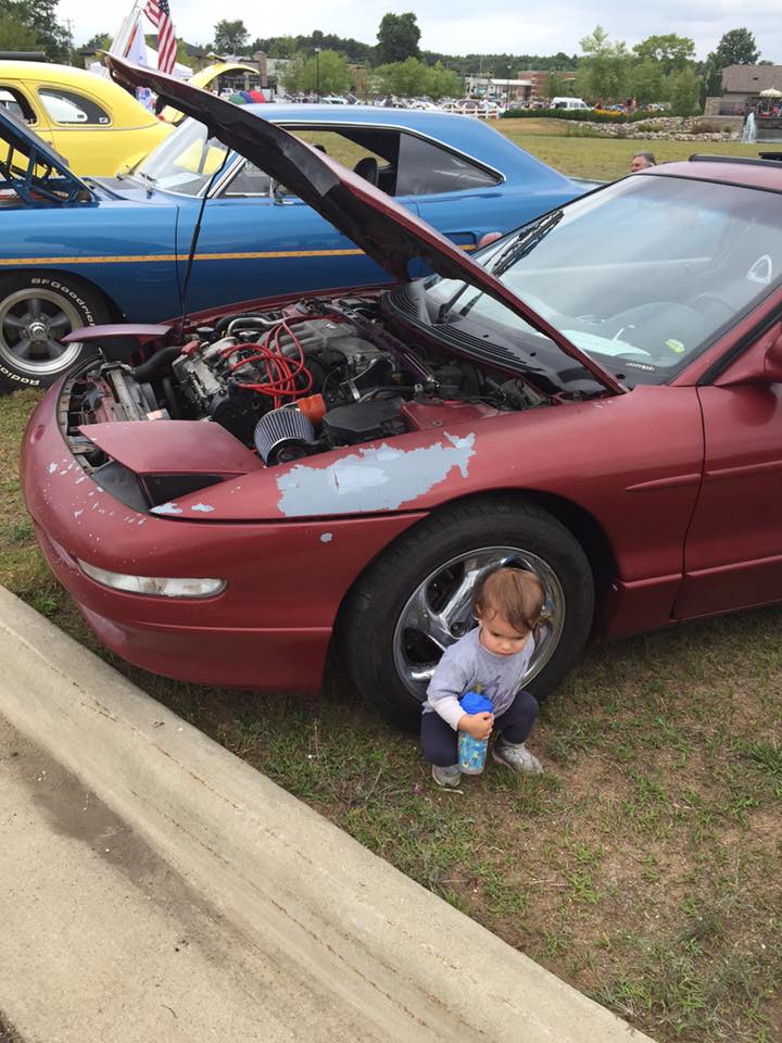 Kid next to a red 1995 Ford Probe with hood open, other cars in background