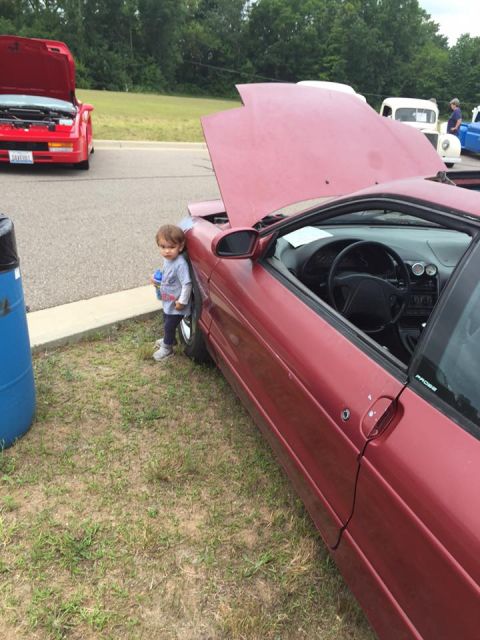1995 Ford Probe - A child stands next to a red car with its hood up, parked on grass.