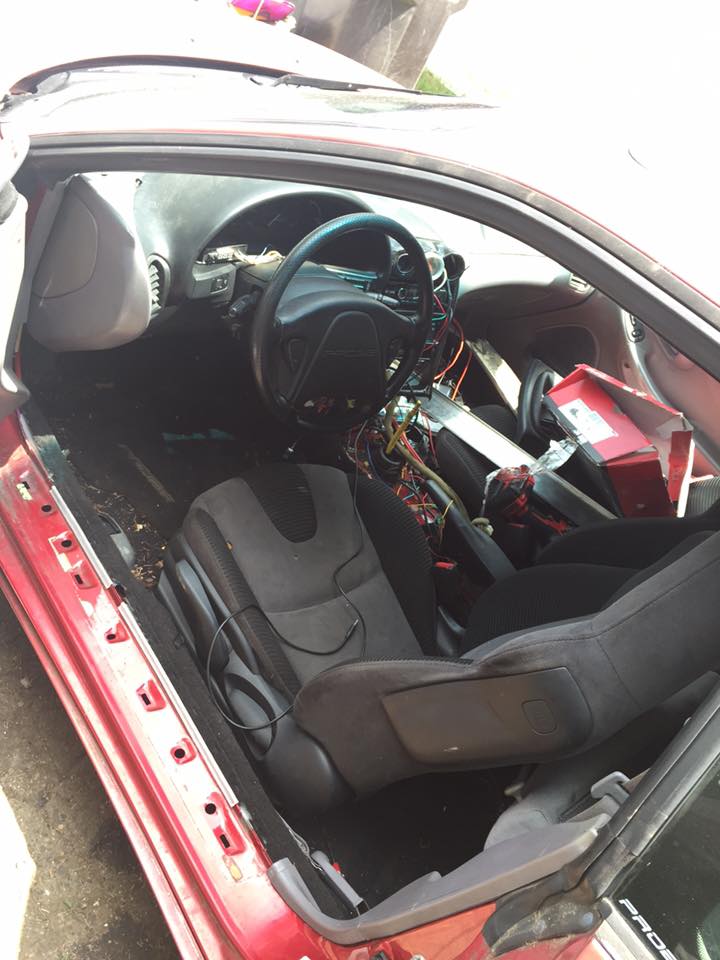 Red car interior with disassembled dashboard, exposed wires, and dark seats. The driver's side door is open