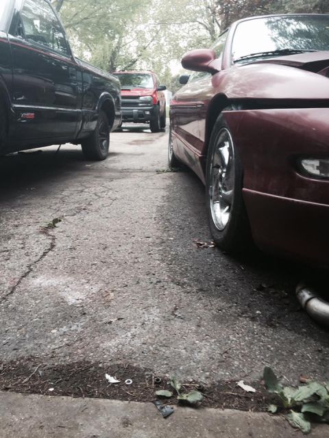 1995 Ford Probe - A low-angle, side view of a red Ford Probe parked on a cracked asphalt driveway next to a black pickup truck. The car's front wheel and par…