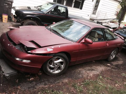 1995 Ford Probe - A red 1990s Ford Probe coupe is parked in a yard with a black pickup truck behind it. The Probe has its hood open, revealing damage to the…