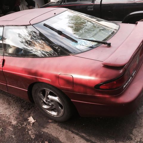 1995 Ford Probe - The rear passenger side of a red 1995 Ford Probe is shown parked outdoors next to a black pickup truck. The car features a rear spoiler and…