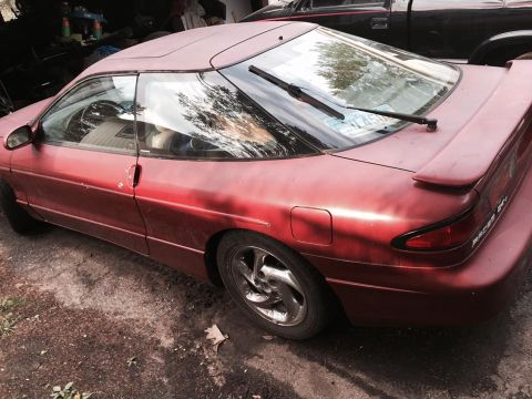1995 Ford Probe - The rear driver's side quarter view of a red 1990s coupe with a rear spoiler. The car is parked on dirt and gravel next to a black truck.