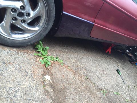 1995 Ford Probe - The lower side panel and wheel of a red car are visible, supported by a red jack stand and a black jack. A screwdriver lies on the concrete…