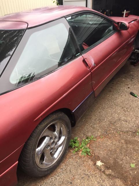 1995 Ford Probe - The right side of a red 1995 Ford Probe is shown parked on a cracked asphalt driveway. The car has a sunroof and a chrome five-spoke wheel…