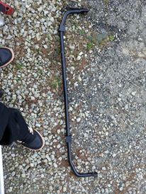 1995 Ford Probe - A black metal sway bar is laid out on a gravel surface. The part is curved and has mounting points on each end.