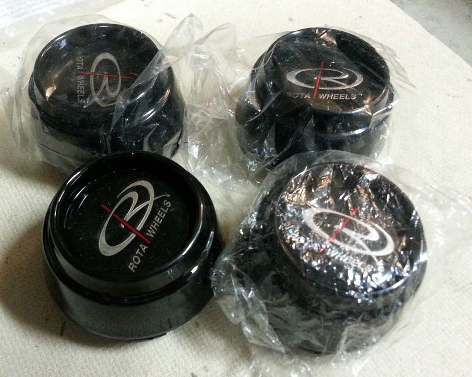 Four black Rota Wheels center caps with silver and red logos, individually wrapped in plastic.