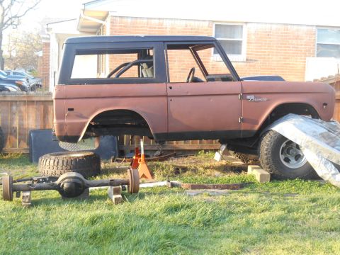 1972 Ford Bronco - A rusty brown 1972 Ford Bronco is elevated on jack stands with its front axle removed and placed on blocks in front of the vehicle. The veh…