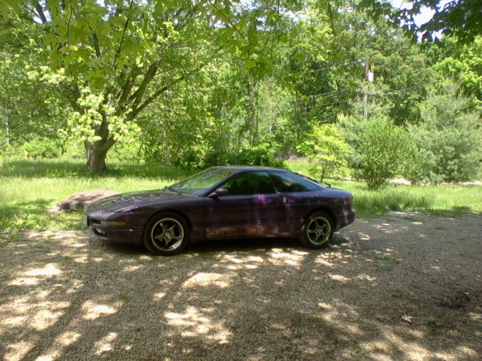 1995 Ford Probe coupe, purple, parked on gravel driveway in dappled sunlight.