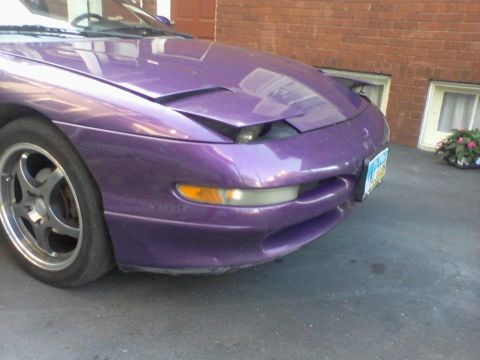 1995 Ford Probe - The front passenger side of a purple 1995 Ford Probe is shown parked on an asphalt driveway in front of a brick building. The car has a pop…
