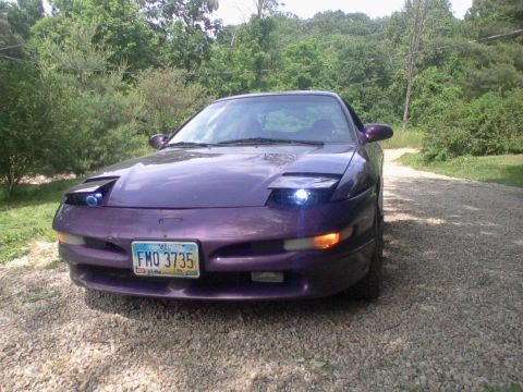 1995 Ford Probe - A purple 1995 Ford Probe is parked on a gravel driveway, facing slightly to the right, with its headlights on. The car is surrounded by lus…