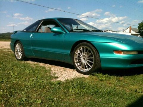 1995 Ford Probe - A side profile of a teal 1995 Ford Probe is parked on a gravel driveway next to a grassy area. The car features aftermarket silver multi-sp…