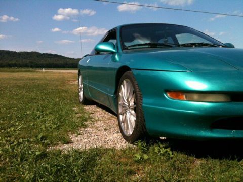 1995 Ford Probe - A teal 1995 Ford Probe is parked on a gravel and grass shoulder next to a field with a tree line in the background.