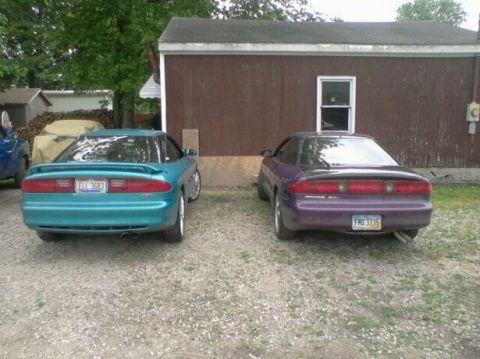 1995 Ford Probe - The rear view of two Ford Probe coupes parked in a gravel lot next to a brown building. The car on the left is teal with a license plate re…