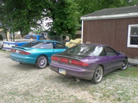 1995 Ford Probe - Two Ford Probe sports coupes are parked on a gravel lot next to a brown building. The teal car is in the foreground left, and the purple ca…