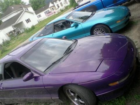 1995 Ford Probe - A purple Ford Probe is parked in front of a teal Ford Probe, with a blue truck visible behind them. The cars are parked on a gravel surface…