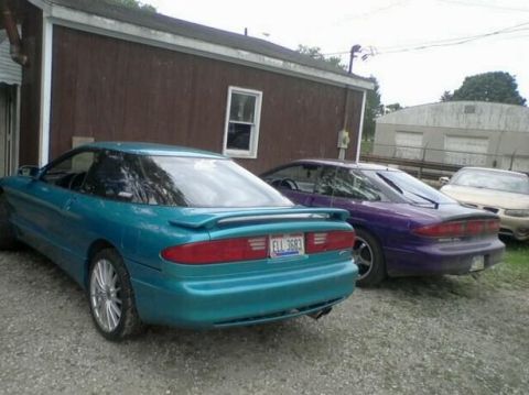 1995 Ford Probe - A teal 1995 Ford Probe is parked next to a purple Ford Probe in front of a brown building. The teal Probe is in the foreground, showing its…