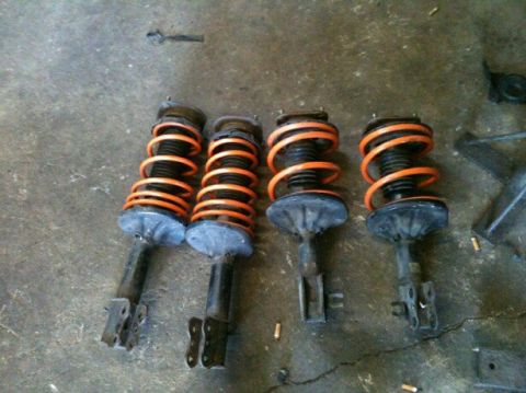 1995 Ford Probe - Four orange coilover suspension assemblies are laid out on a concrete floor. Each assembly consists of an orange coil spring over a black s…