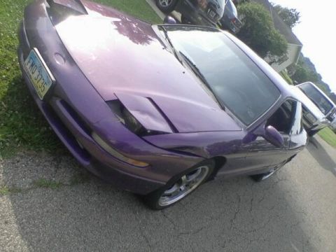 1995 Ford Probe - A low-angle, slightly tilted shot of a purple Ford Probe parked on an asphalt driveway next to a grassy area. The front passenger side of t…