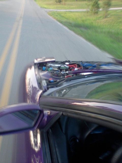 1995 Ford Probe - A first-person perspective shot from inside a purple car driving down a road. The hood is open, revealing the engine components and wiring.